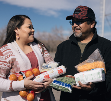 A couple seeking help have their arms full of nutritious foods while smiling at each other.