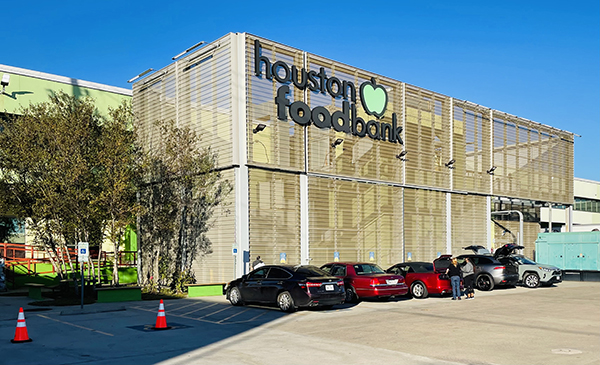 Under a blue sky sits the industrial-looking building with the large words Houston Food Bank attached to the side. Cars parked along the building awaiting their grocery pick up.