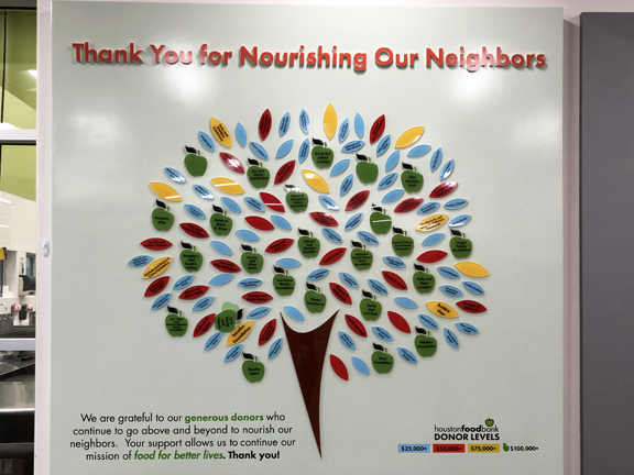 A replica of a tree, made from acrylic, has leaves of yellow, blue and red along with large green apples. Several of the leaves and apples contain names of corporate and individual donors who have made gifts to the Food Bank.