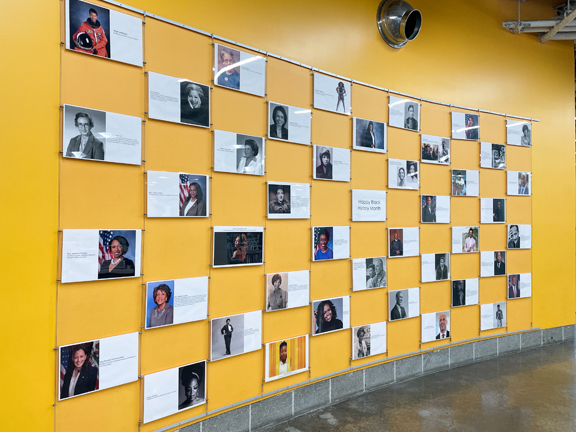 img_tour_photowall A bright yellow wall with rows of frames featuring rows of photos and descriptions of prominent African American icons during Black History Month.