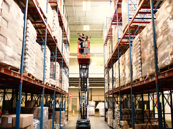 img_tour_warehouse A scissor lift expanded high in the air between two warehouse rafters full of food pallets.