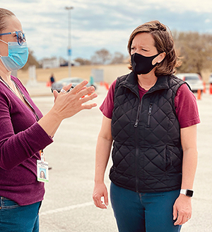 Two masked women stand in large parking lot at a drive through food distribution discussing the issues of hunger and the importance of food banks.