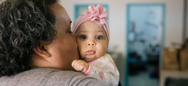 A mother stands in her home embracing her young infant daughter with a kiss on her cheek. 