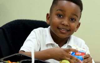 A child sits at a table opening a small carton of milk with a tray of food in front of him.