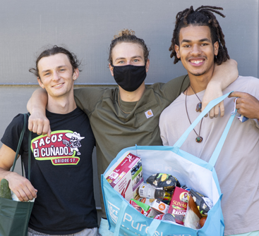 Three college students stand together posing for the camera holding up a large blue bad of grocery products including fresh fruits and vegetables.