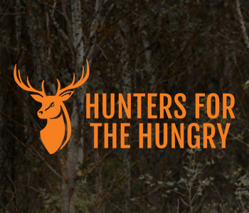 A darkly lit wooded area with trees and brush with the words "Hunters For The Hungry" in bright orange.