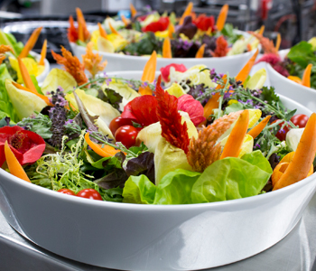 A brightly colorful salad with fresh greens, carrots and other vegetables artistically sit in a large white, glass bowl.