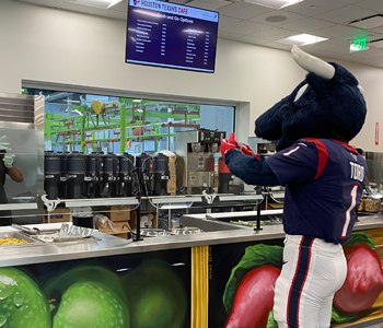 Toro, the Houston Texans mascot, stands in a cafeteria lined food service in the Texans Cafe at Houston Food Bank.